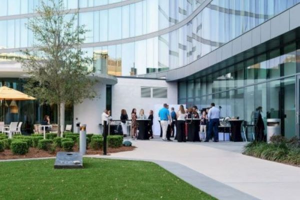 People are gathered outside a modern glass building, likely at a social event. Tables and umbrellas are set up.