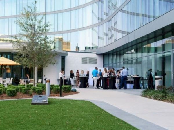 People are gathered outside a modern glass building, likely at a social event. Tables and umbrellas are set up.