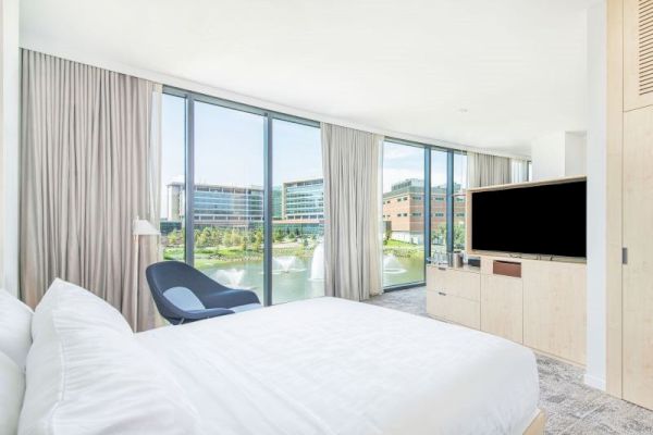 A modern bedroom with a large bed, a TV on a cabinet, an armchair, and floor-to-ceiling windows offering a view of buildings and greenery.