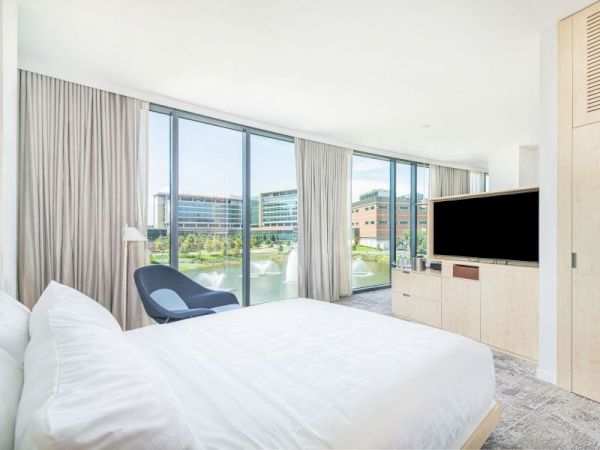 A modern bedroom with a large bed, a TV on a cabinet, an armchair, and floor-to-ceiling windows offering a view of buildings and greenery.