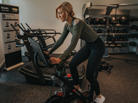 A person in athletic wear is using a stationary bike in a gym setting, surrounded by fitness equipment and weights on shelves, focused on the workout.