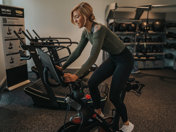 A person in athletic wear is using a stationary bike in a gym setting, surrounded by fitness equipment and weights on shelves, focused on the workout.