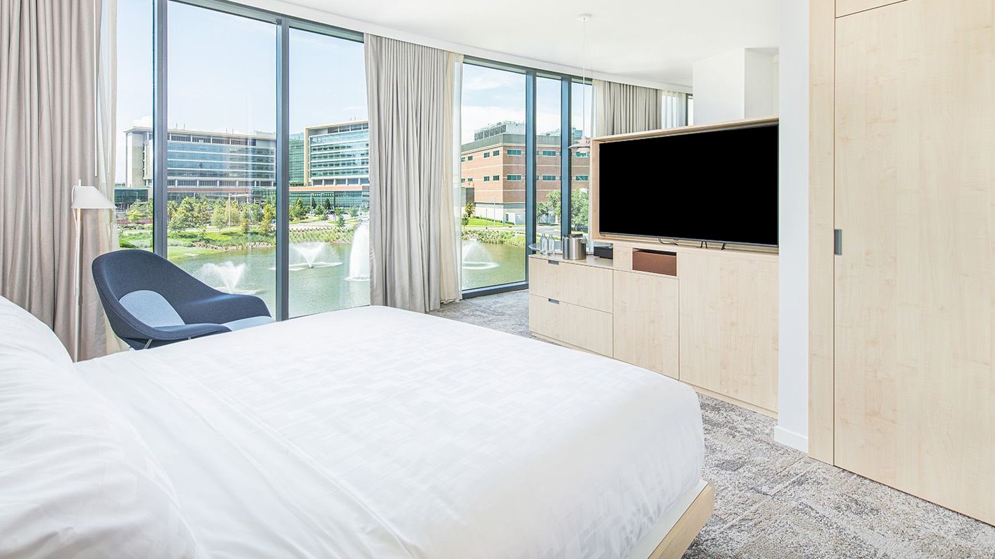 This image shows a modern hotel room with a large bed, a chair, a TV, and a scenic view outside the floor-to-ceiling windows ending the sentence.
