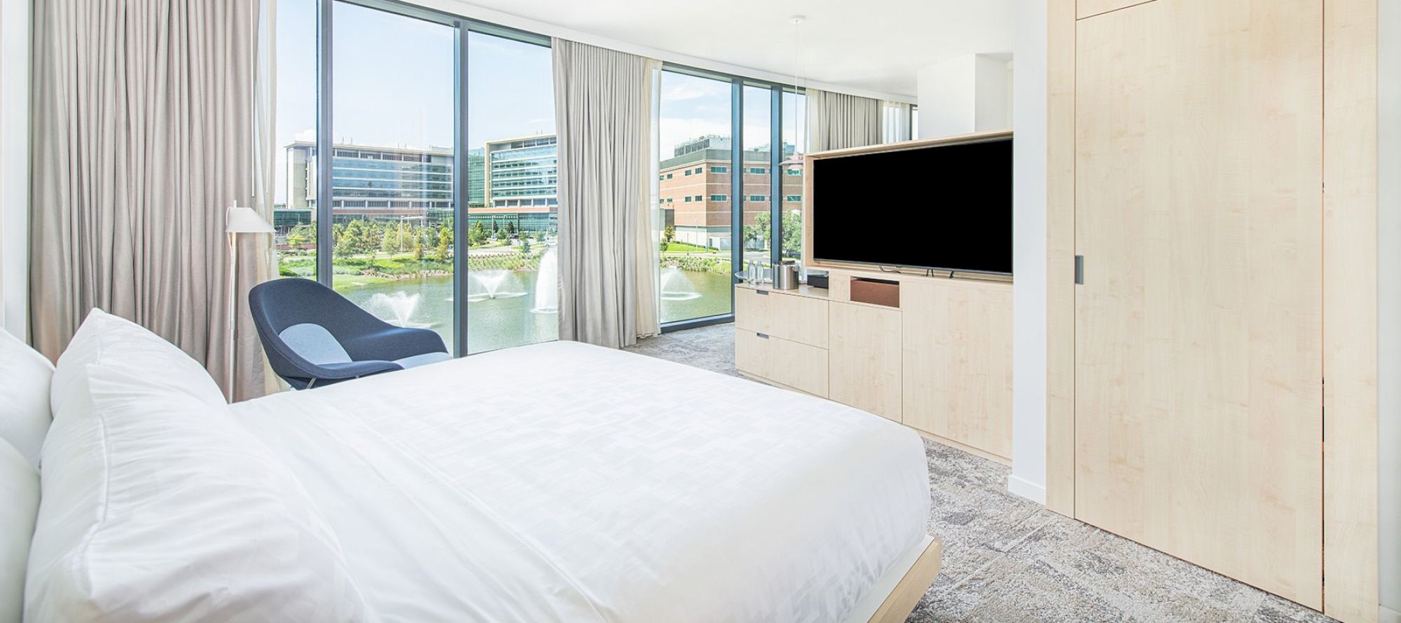 This image shows a modern hotel room with a large bed, a chair, a TV, and a scenic view outside the floor-to-ceiling windows ending the sentence.