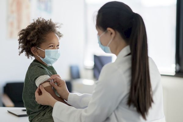 A healthcare provider wearing a mask is preparing a young child, also masked, for a medical procedure or vaccination in a clinic setting.