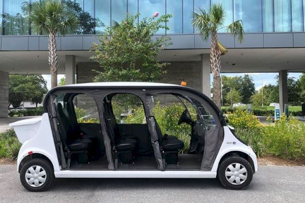 The image shows a white, open-sided electric shuttle parked in front of a modern building with glass windows and flanked by palm trees.