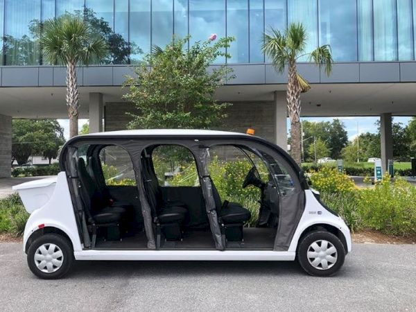 The image shows a white, open-sided electric shuttle parked in front of a modern building with glass windows and flanked by palm trees.