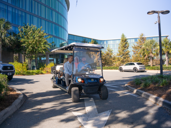 A small golf cart-style vehicle with two people riding inside, driving through a sunny parking lot near a modern glass office building.