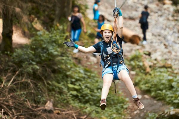 A person wearing a helmet and safety gear is joyfully zip-lining through a forested area, with others visible in the background on a trail.