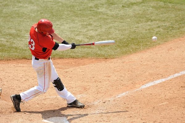 A baseball player wearing a red jersey and helmet, number 23, swings a bat and hits the ball on a baseball field during a game.