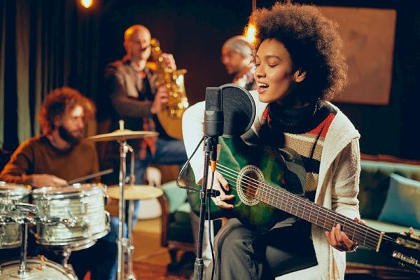 A group of musicians playing instruments in a studio, with a woman singing and playing the guitar at the forefront and others on drums and saxophone.