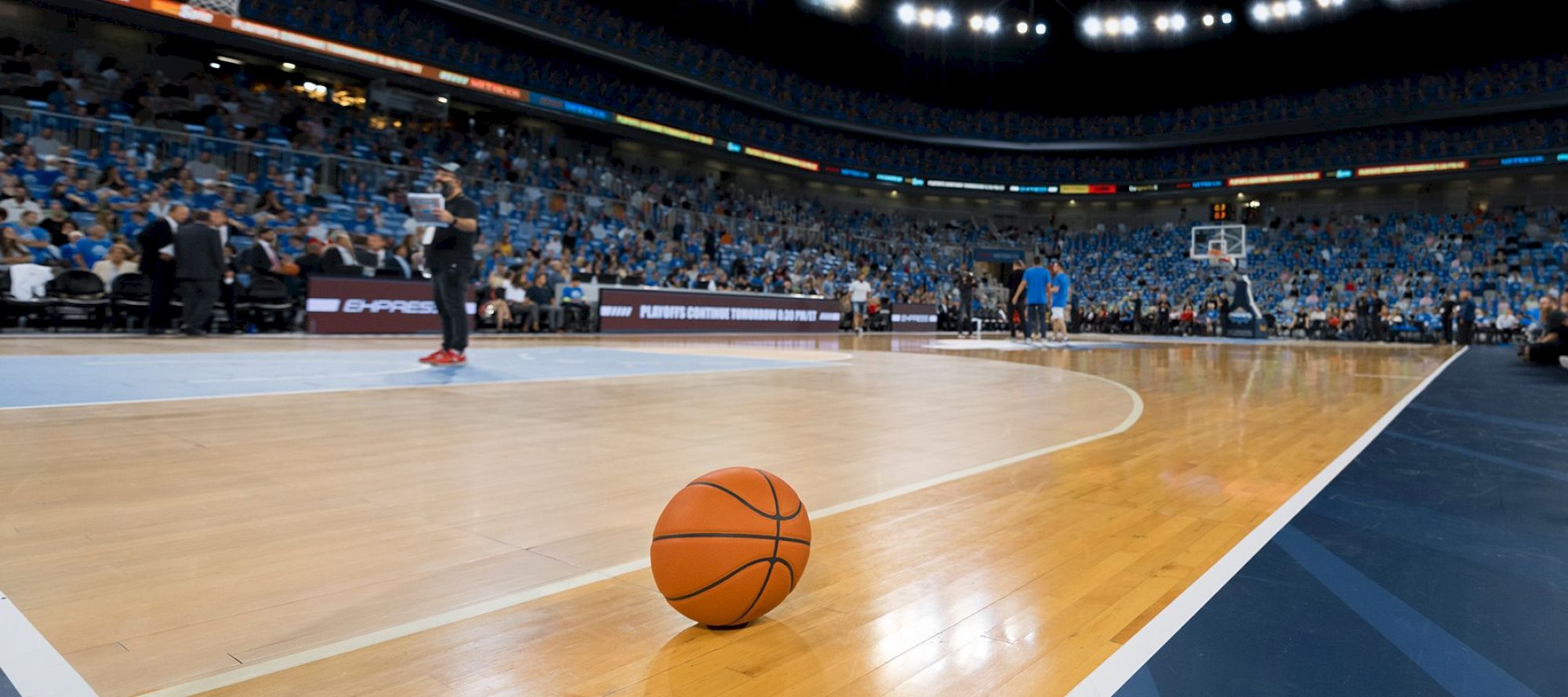 The image shows a basketball on an indoor basketball court with players and spectators in the background during a game or practice.