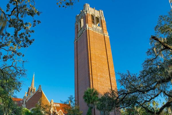 The image shows a tall, red-brick clock tower against a clear blue sky, surrounded by trees and buildings with pointed roofs.