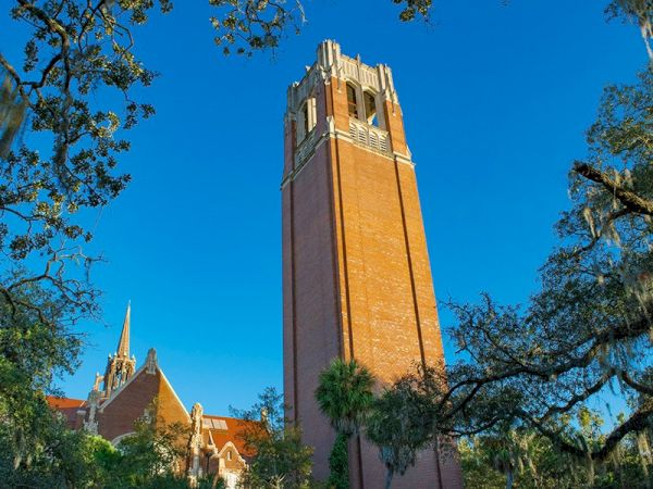 The image shows a tall, red-brick clock tower against a clear blue sky, surrounded by trees and buildings with pointed roofs.