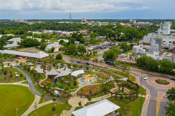 The image shows an aerial view of a park with paths, green spaces, playgrounds, and nearby buildings, under a cloudy sky.