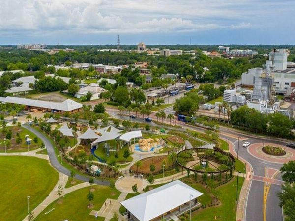 The image shows an aerial view of a park with paths, green spaces, playgrounds, and nearby buildings, under a cloudy sky.