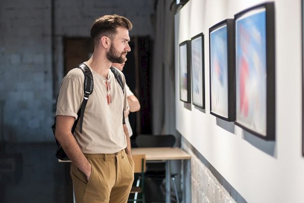 A person with a backpack observes framed artwork on a gallery wall, standing with hands in pockets in a well-lit room.