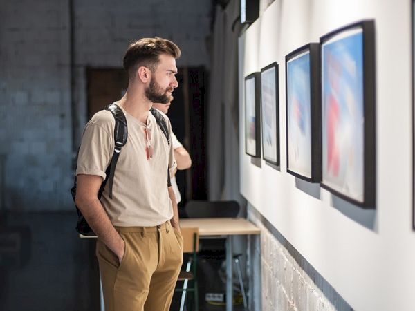 A person with a backpack observes framed artwork on a gallery wall, standing with hands in pockets in a well-lit room.