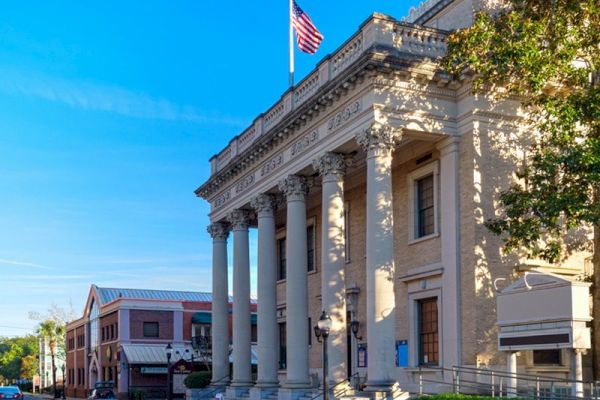 The image shows a historic building with classical columns and an American flag on top, located on a street with other buildings and trees.