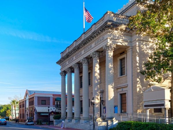 The image shows a historic building with classical columns and an American flag on top, located on a street with other buildings and trees.