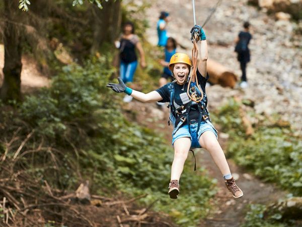 A person wearing safety gear and a helmet is zip-lining in a forested area, with other people visible in the background.