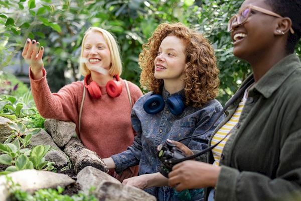 Three people enjoying a lush, green environment; two have headphones around their necks, and one holds a camera, all smiling and interacting.