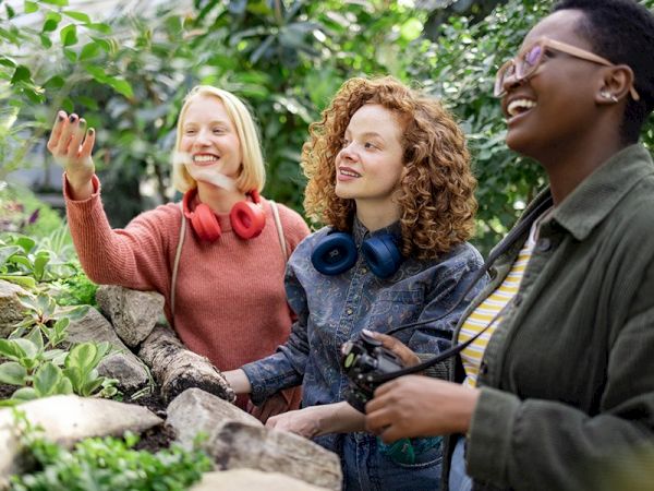 Three people enjoying a lush, green environment; two have headphones around their necks, and one holds a camera, all smiling and interacting.
