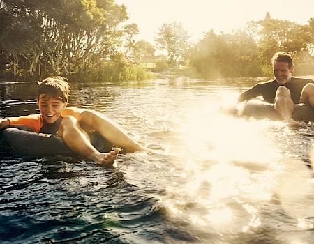 Two people are joyfully floating on inflatable tubes in a sunlit river, surrounded by greenery under a bright, warm sunshine.