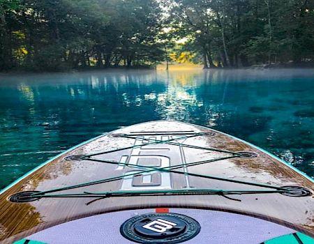 The image shows the front view of a paddleboard on clear blue water, with a misty forest in the background and sunlight streaming through the trees.