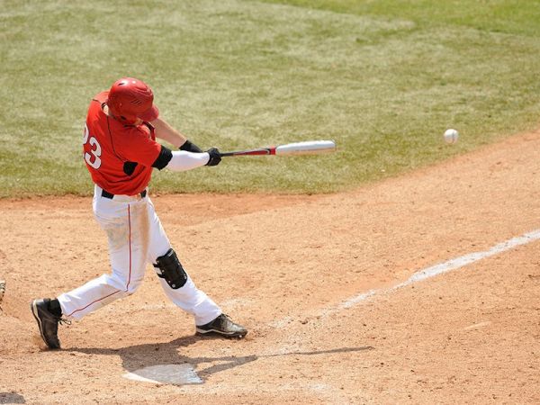 A baseball player in a red jersey and helmet, wearing number 23, makes contact with a pitched ball on a dirt baseball field, ending the sentence.