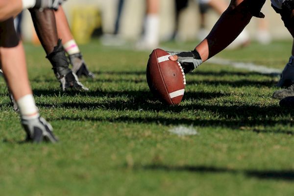 The image shows a close-up of American football players at the line of scrimmage, with one player holding the football on the ground.