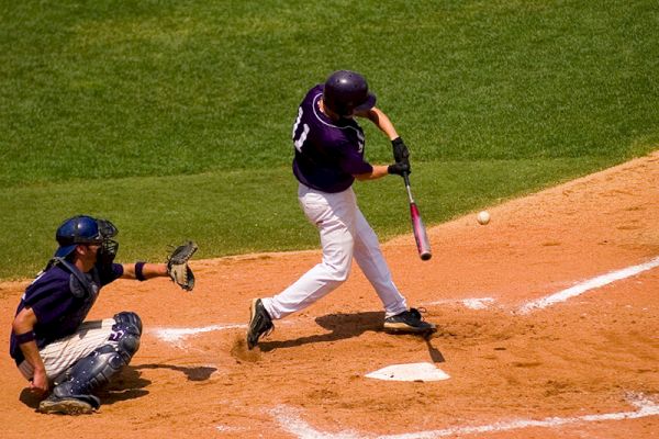 A baseball player wearing a dark jersey is hitting the ball while a catcher crouches behind home plate. The scene takes place on a sunny field.