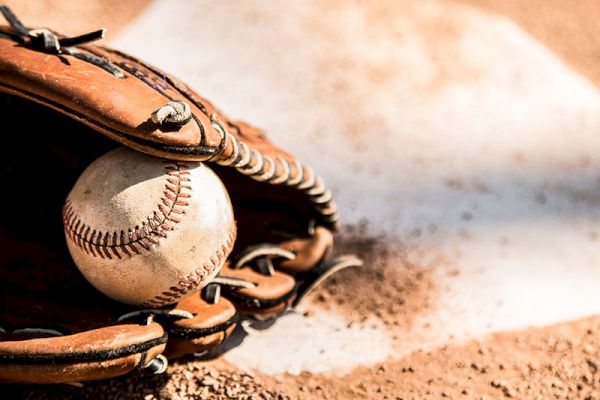 A baseball glove holding a baseball, placed on the ground near home plate on a baseball field, with some dirt visible.