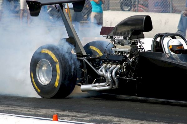 A drag racing car is performing a burnout, generating smoke from its rear tires at a racetrack with a crowd visible behind a fence.