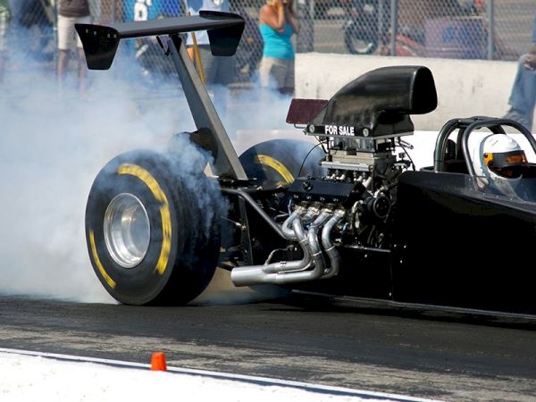 A drag racing car is performing a burnout, generating smoke from its rear tires at a racetrack with a crowd visible behind a fence.
