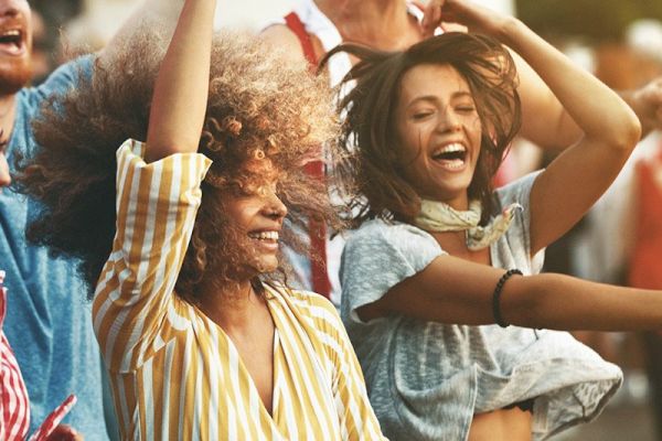 Two women are joyfully dancing in a lively outdoor setting, surrounded by other people.