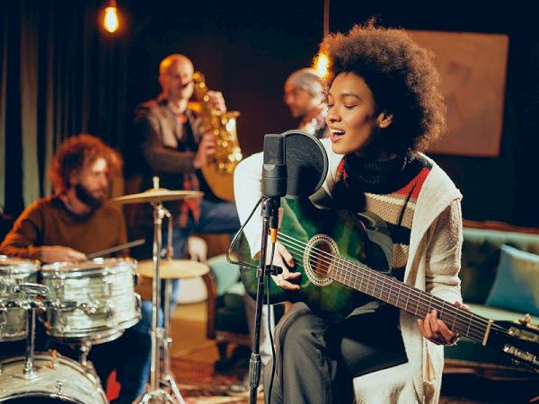 A group of musicians is performing in a cozy room with dim lighting, featuring a woman singing and playing guitar, a drummer, and a saxophonist.