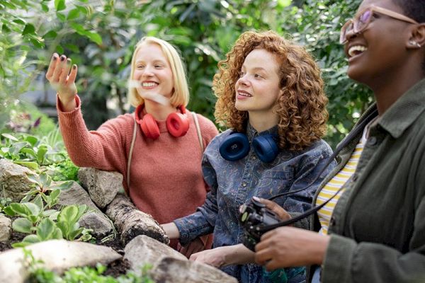 Three people are enjoying a garden, looking at plants and smiling, with two wearing headphones and one holding a camera.