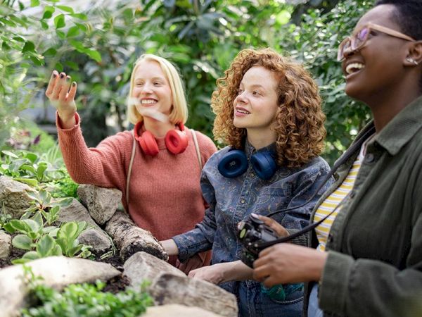 Three people are enjoying a garden, looking at plants and smiling, with two wearing headphones and one holding a camera.