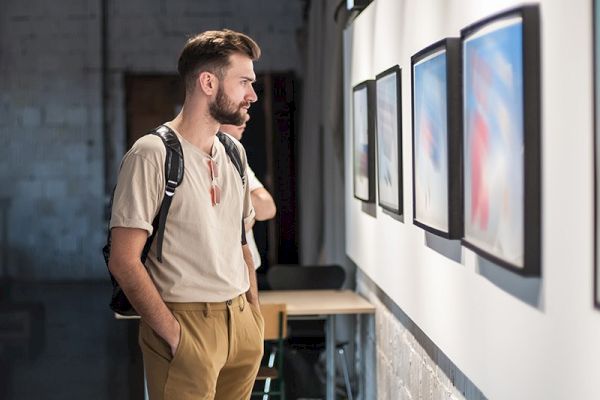 A man with a backpack is intently looking at framed artworks on a gallery wall.