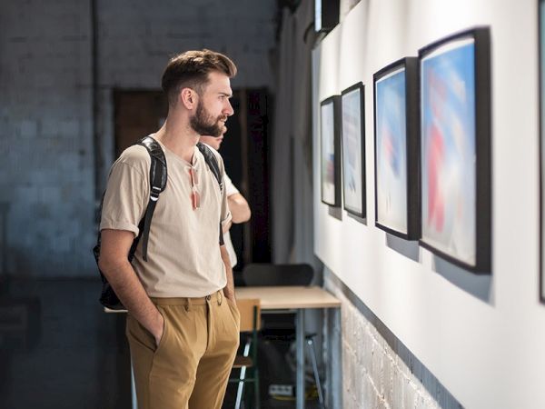 A man with a backpack is intently looking at framed artworks on a gallery wall.