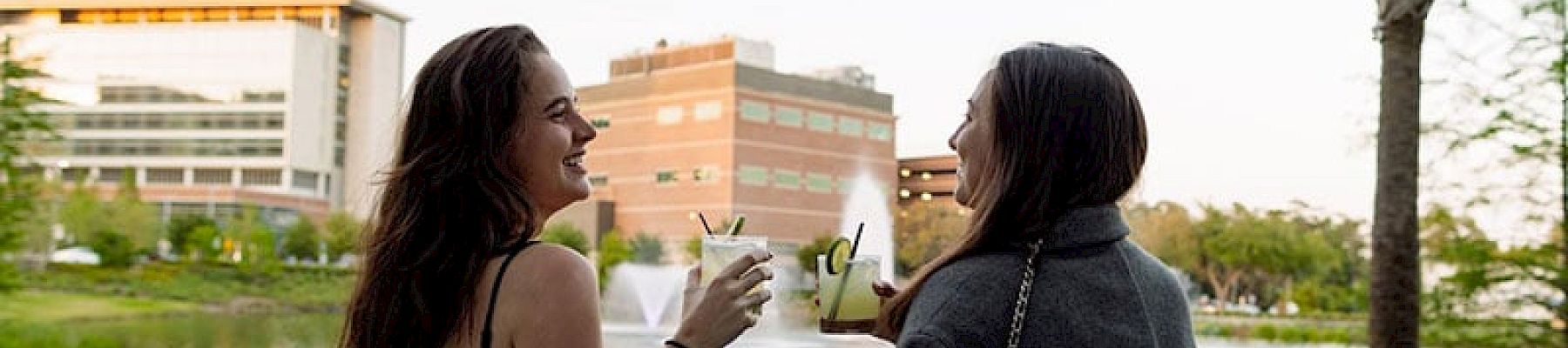 Two people are sitting on a sofa outdoors, enjoying drinks and having a conversation, with buildings and a palm tree in the background.