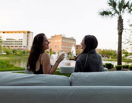 Two people are sitting on a sofa outdoors, enjoying drinks and having a conversation, with buildings and a palm tree in the background.