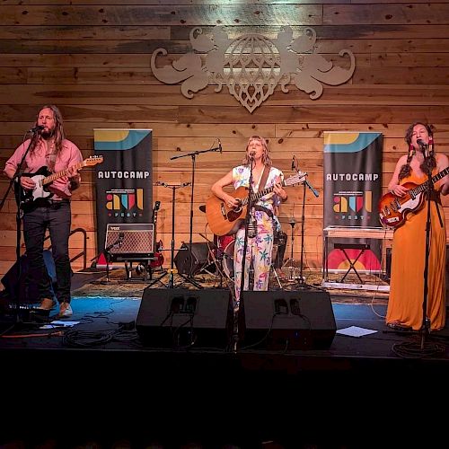 Three musicians perform on a stage with a wooden backdrop, flanked by banners that read 