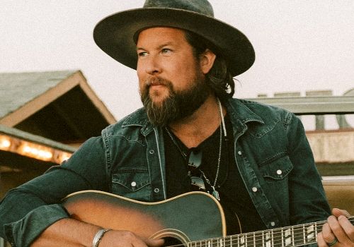 A bearded man wearing a hat and denim jacket is playing an acoustic guitar outdoors. He appears focused and engaged with his music.