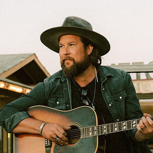 A bearded man wearing a hat and denim jacket is playing an acoustic guitar outdoors. He appears focused and engaged with his music.