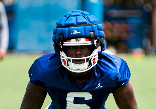 A football player wearing a blue padded helmet and jersey number 6, white pants, and blue gloves, is in a ready stance on the field.