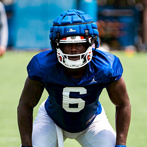 A football player wearing a blue padded helmet and jersey number 6, white pants, and blue gloves, is in a ready stance on the field.