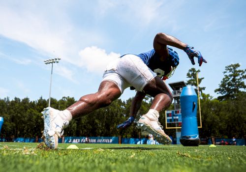 An athlete in football gear is performing a practice drill on a grassy field, with practice equipment visible and trees in the background.