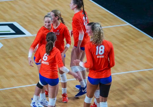 A group of female volleyball players wearing orange jerseys stand together on an indoor court, seemingly celebrating a point scored.